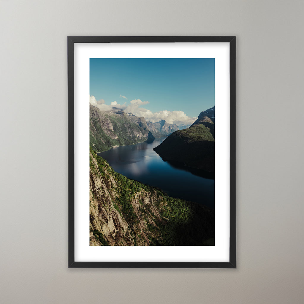 Dramatisk utsikt över en djupblå fjord i Norge, omgiven av höga, grönskogade berg och klippväggar under en delvis molnig himmel i olika storlekar: 30x40 cm, 40x50 cm, 50x70-cm, 70x100 cm.