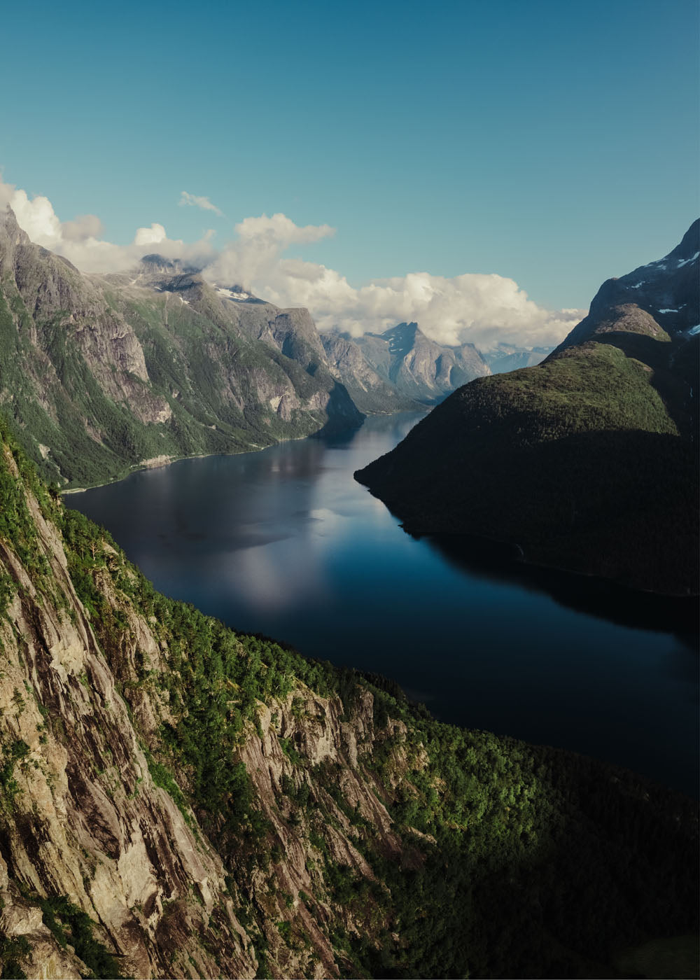 Dramatisk utsikt över en djupblå fjord i Norge, omgiven av höga, grönskogade berg och klippväggar under en delvis molnig himmel i olika storlekar: 30x40 cm, 40x50 cm, 50x70-cm, 70x100 cm.