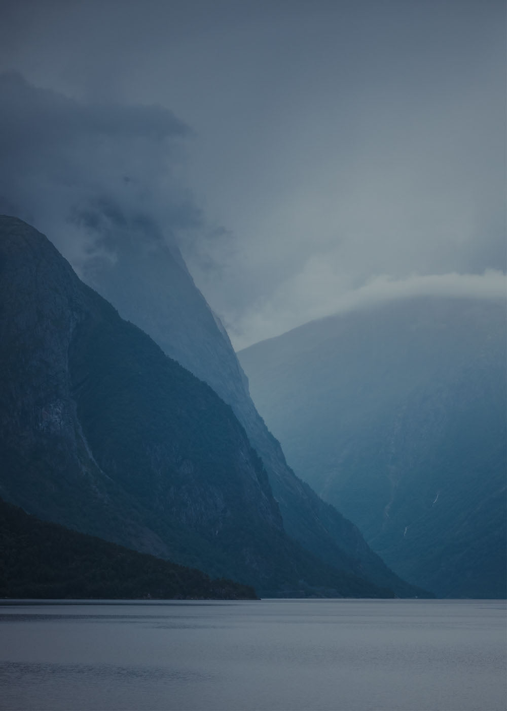 Dimmig fjordlandskap med höga blå berg som reser sig från lugnt vatten. Poster i olika storlekar: 30x40 cm, 40x50 cm, 50x70-cm, 70x100 cm.